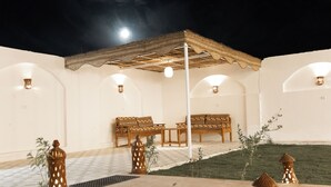 Un espacio al aire libre techado con mesas y bancos, rodeado de muros con ventanas de arco y luces fijadas en la pared.