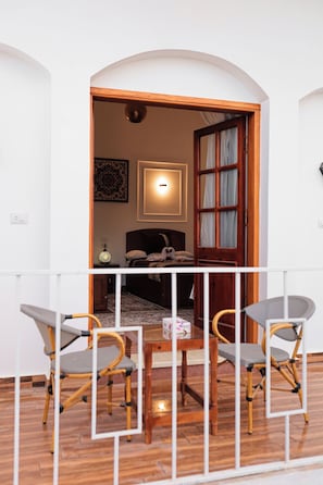 Un balcón con una mesa y sillas de madera, vista hacia un cuarto con cama y lámpara.