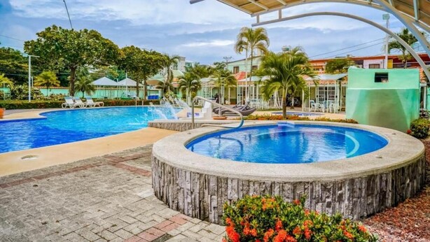 Piscina com deck de madeira e espreguiçadeiras, cercada por palmeiras e uma área coberta.
