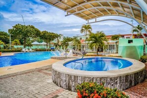 A swimming pool with a wooden deck and lounge chairs, surrounded by palm trees and a covered area. - Fabulous 3-bedroom condo with AC in vibrant Jaco (Jaco)