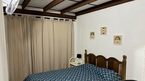Quarto com cama, cadeira, mesa de cabeceira e ventilador.