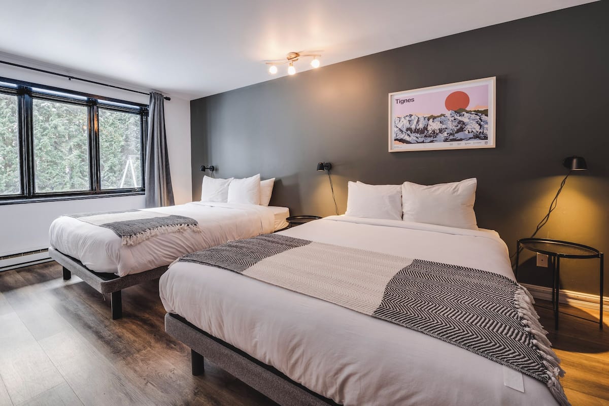A modern bedroom with two beds, a large window, a wall-mounted artwork, and a bedside table with a lamp.