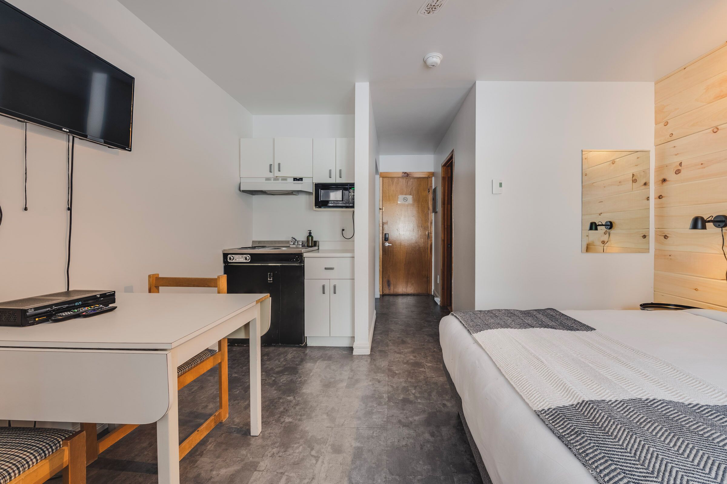 Una habitación de hotel compacta con cama, escritorio, televisor, kitchenette y una pared de acento de madera.