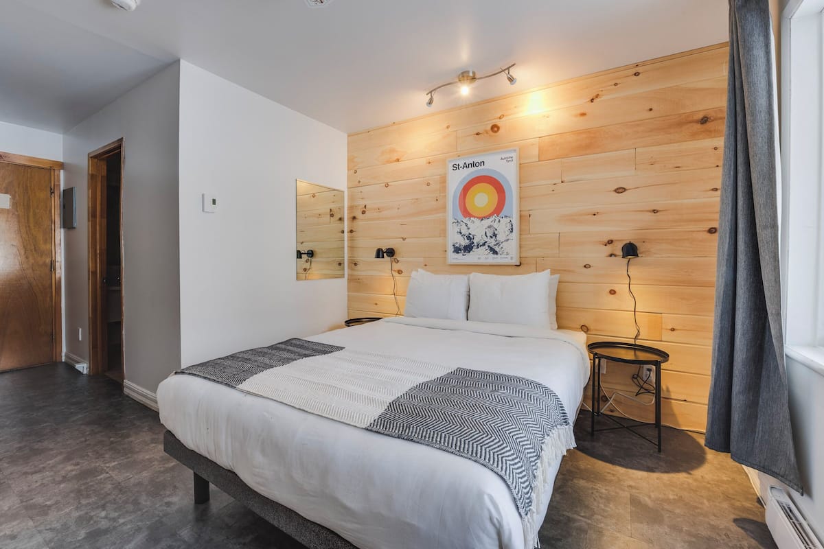 A bedroom with a bed, a nightstand, a wall-mounted light, a poster, and a wooden wall.