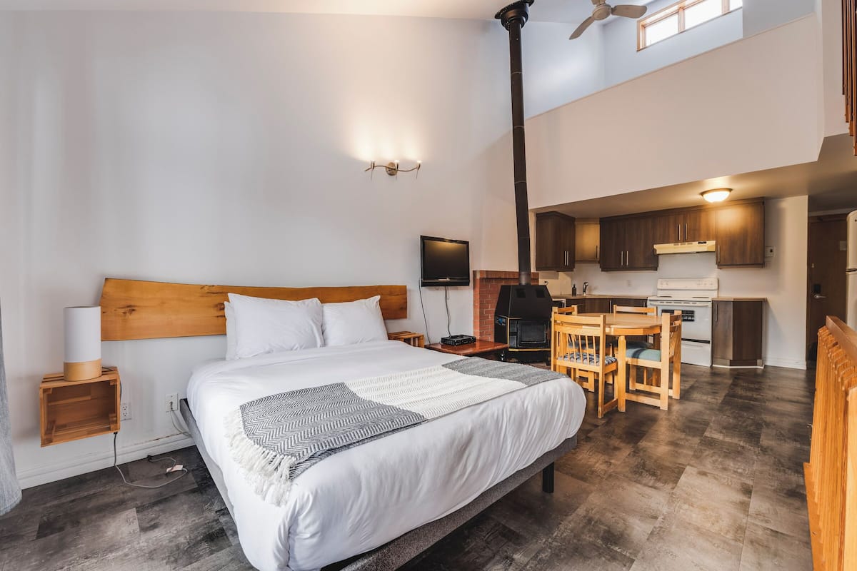 A bedroom with a large bed, bedside table, lamp, and a view of the kitchen area.