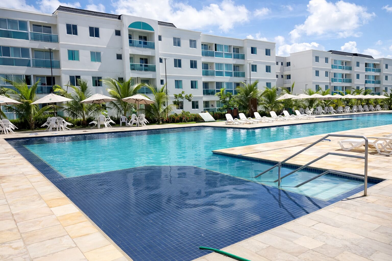 Une piscine entourée de chaises longues et de parasols, bordée d’un immeuble doté de nombreux balcons, sous un ciel dégagé.