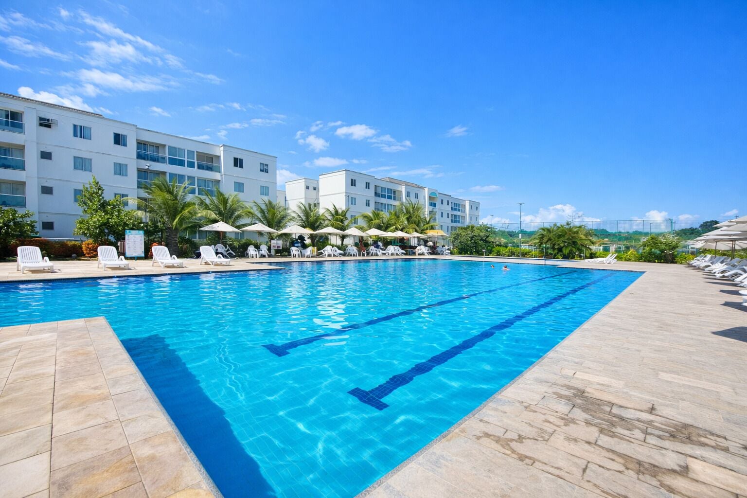 A swimming pool with clear blue water, surrounded by lounge chairs and umbrellas, with a building in the background.