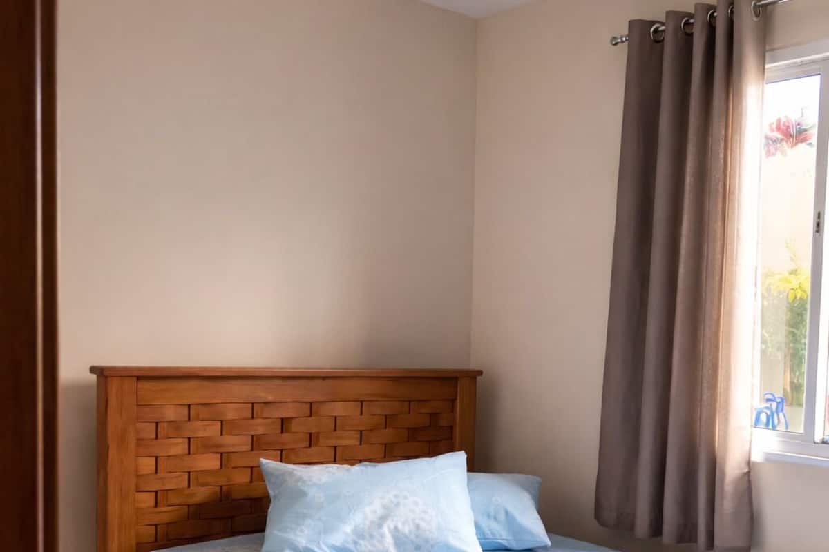 A bedroom with a wooden headboard, a bed with a blue patterned bedspread, a single pillow, and a window with curtains.