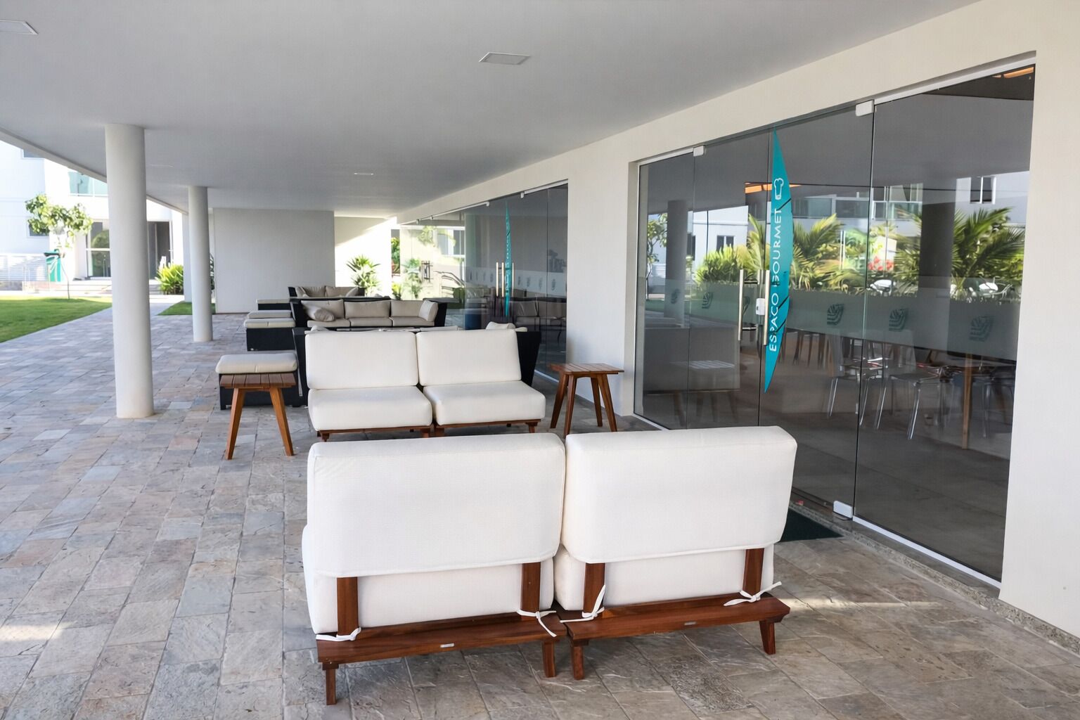 A covered outdoor seating area with white cushioned chairs and wooden tables, large glass doors, and a view of the outdoors.