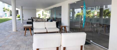 A covered outdoor seating area with white cushioned chairs and wooden tables, large glass doors, and a view of the outdoors.
