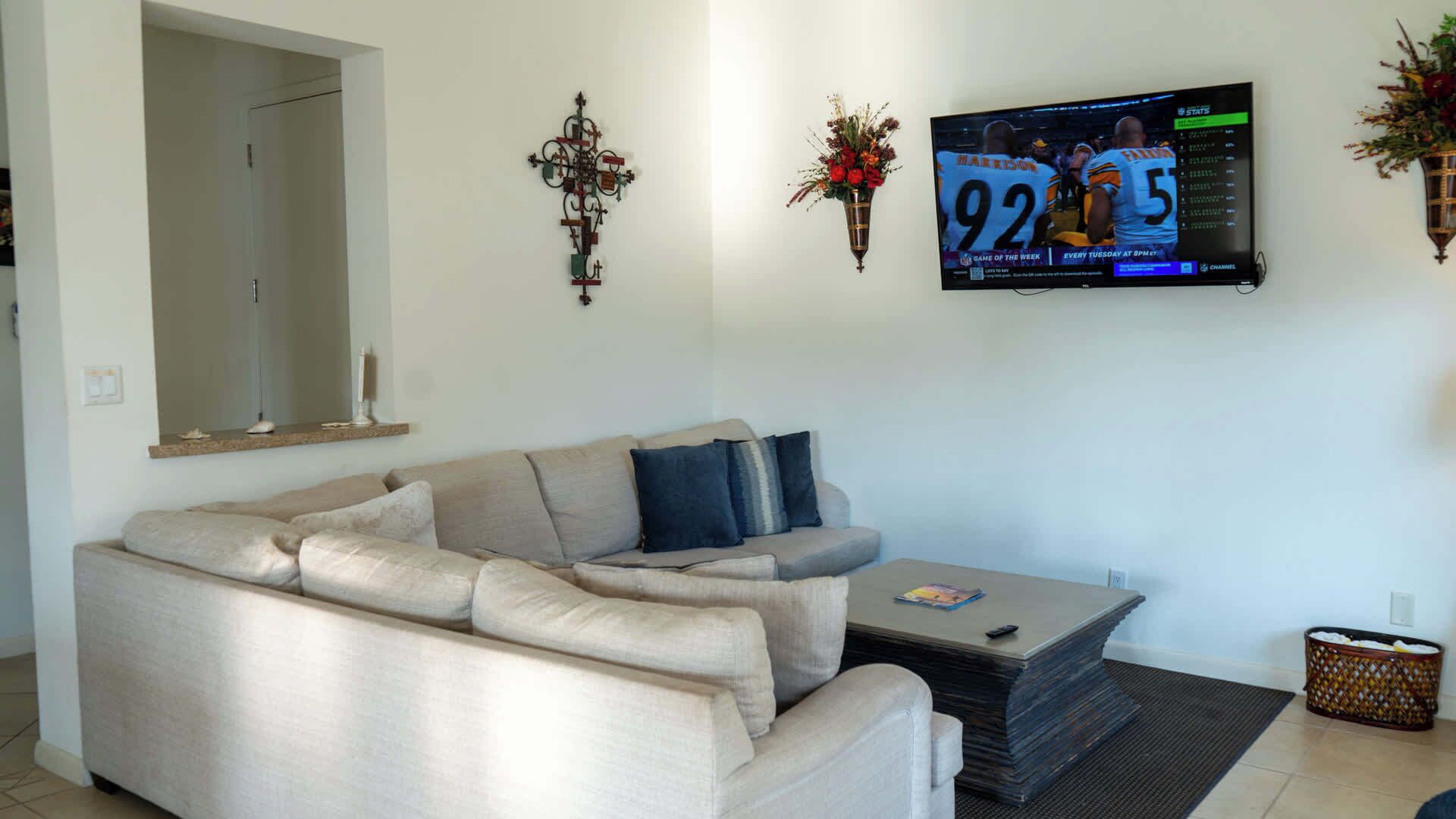 A living room with a beige sectional sofa, a coffee table, a flat-screen TV mounted on the wall, and a decorative wall piece.