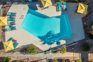 Ovenudblik over en swimmingpool med liggestole og parasoller, omgivet af en belagt terrasse og palmer.