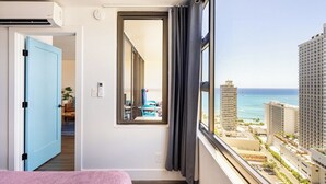 A hotel room with a bed, a window with a view of the ocean and buildings, a door, and a small mirror. - Waikiki Banyan Ocean View Suite, High Floor Remodeled, AC & Free Parking (Honolulu)