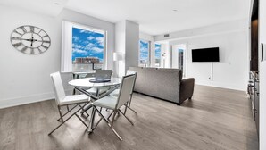 Une chambre d’hôtel moderne dotée d’un coin repas, d’une grande fenêtre offrant une vue sur la ville, d’un téléviseur à écran plat et d’une horloge murale.