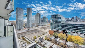 Un immeuble de grande hauteur doté d’une façade moderne en verre, une voie ferrée qui longe le bâtiment, et un mélange de bâtiments résidentiels et commerciaux en arrière-plan.