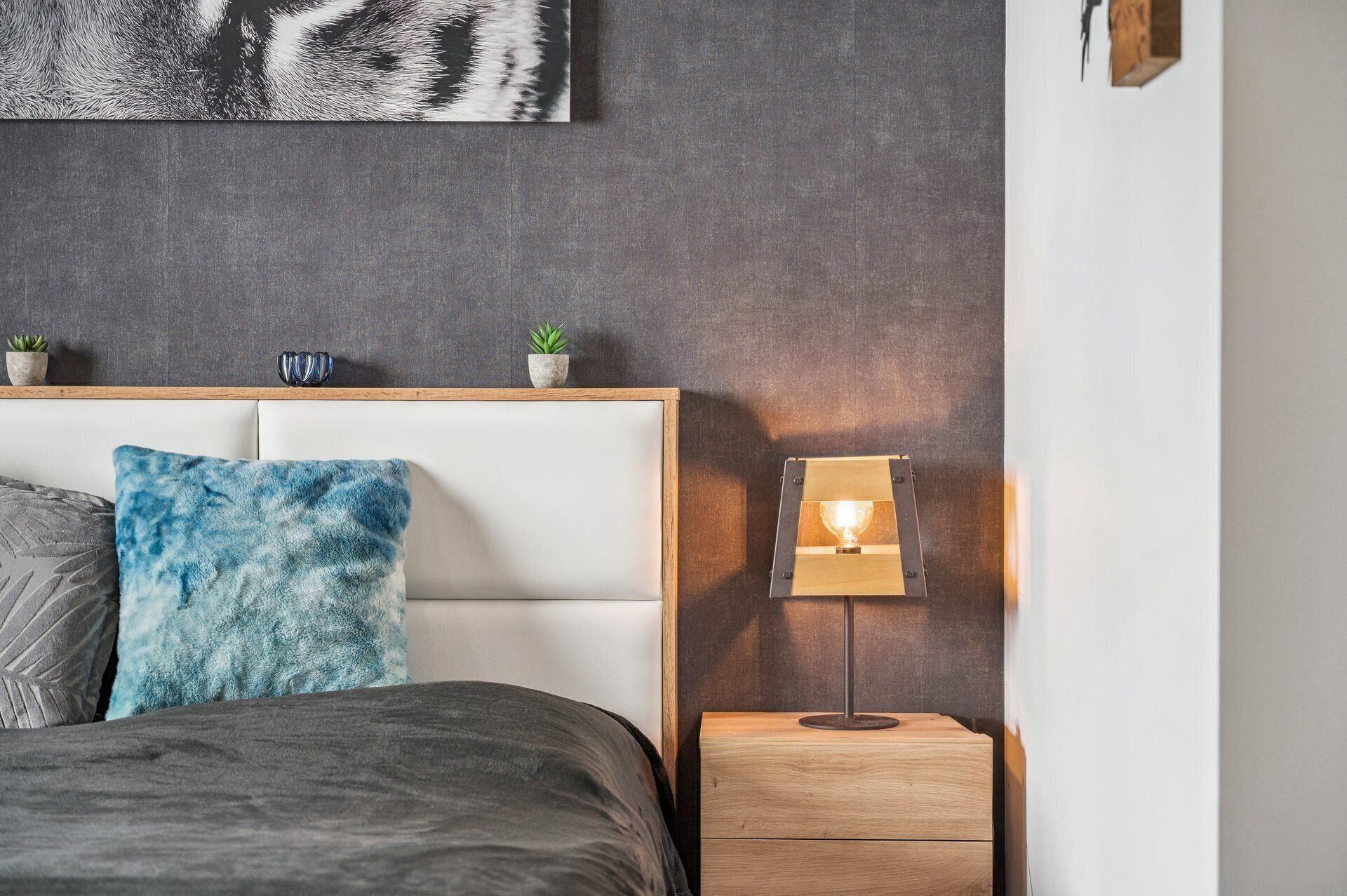 A bedroom with a bed, a nightstand with a lamp, a wall-mounted shelf with plants, and a large abstract painting.