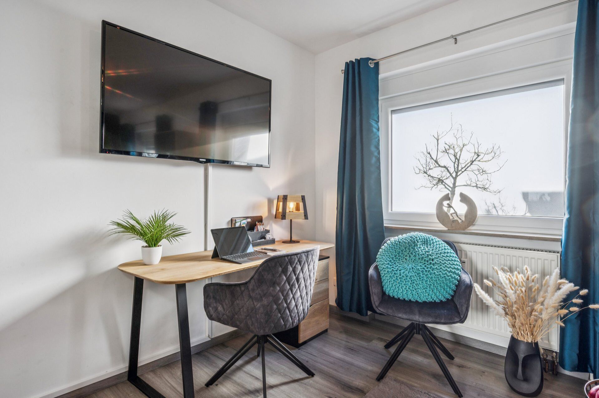 Ein modernes Zimmer mit Schreibtisch, Stuhl, Fernseher und einem Fenster mit Vorhängen.