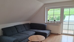 Ein modernes Wohnzimmer mit einem Ecksofa, einem kleinen Holztisch und einem großen Fenster mit Blick auf Grünflächen.