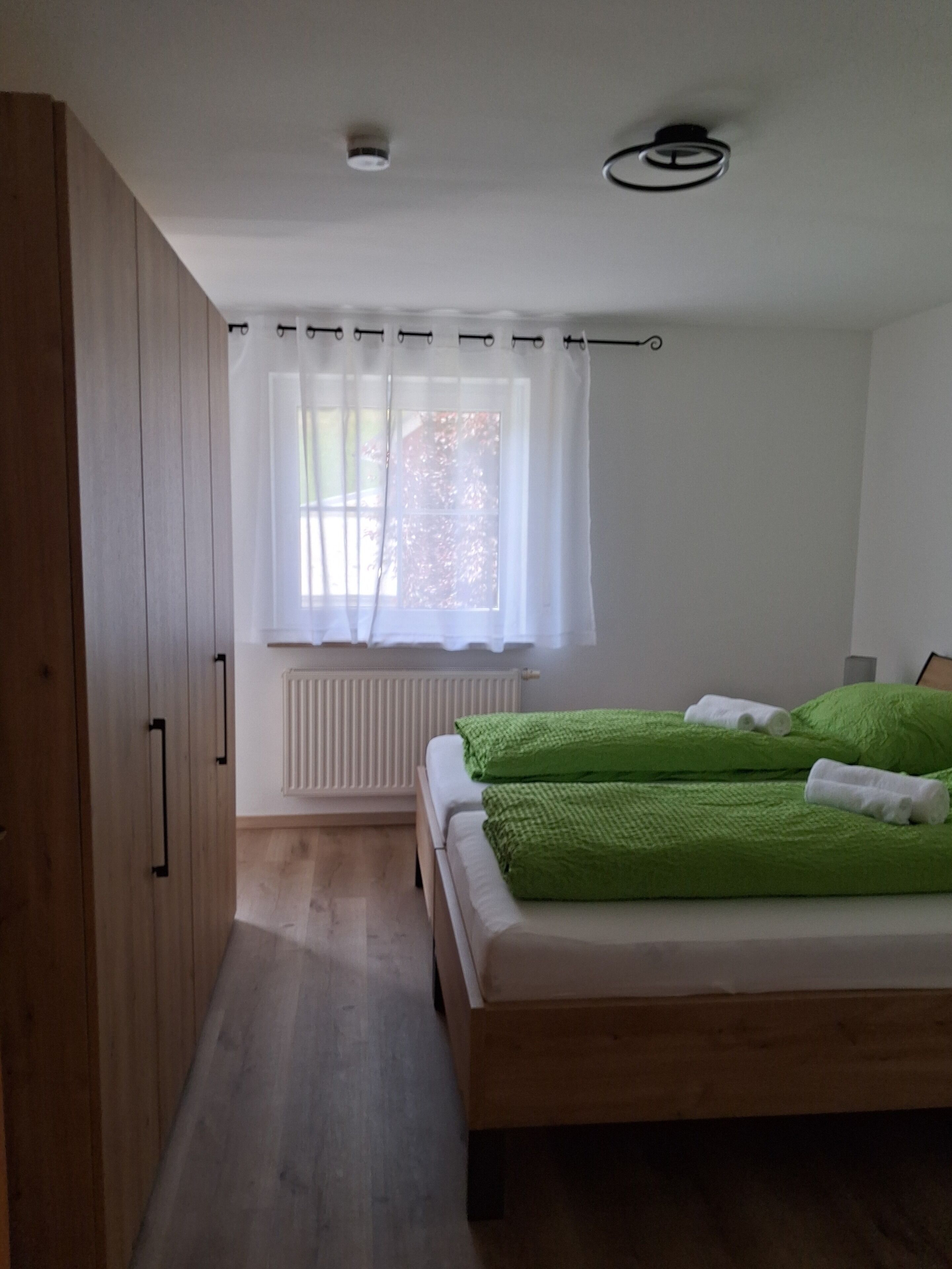 Ein Schlafzimmer mit einem Holzschrank, einem Fenster mit weißen Vorhängen, einem Heizkörper und einem Bett mit grüner Bettwäsche.