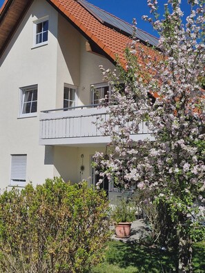 Un edificio de dos pisos con un balcón, un árbol de flores y una planta en maceta.