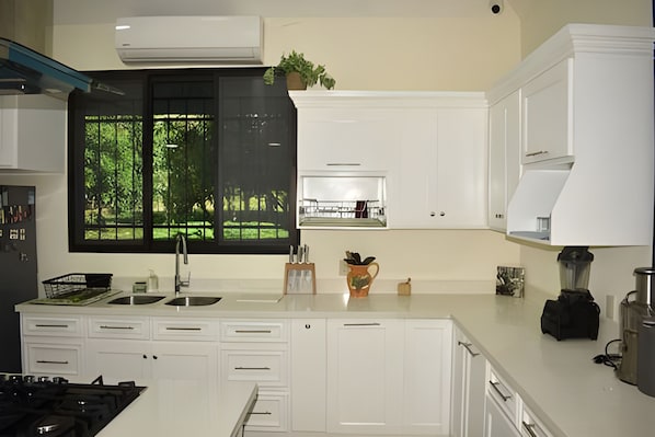 Une cuisine moderne avec des meubles blancs, une grande fenêtre donnant sur un espace verdoyant, un évier et une cuisinière.