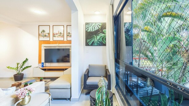 A modern living room with a flat-screen TV, comfortable seating, a dining area, and a large window with a view of palm trees. - Complete Apartments in Asa Norte - BSB BVT (Brasília)