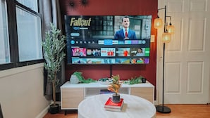 Un salon avec un téléviseur à écran plat diffusant une émission, une table basse ronde sur laquelle se trouve un livre, une plante en pot et une lampe moderne.