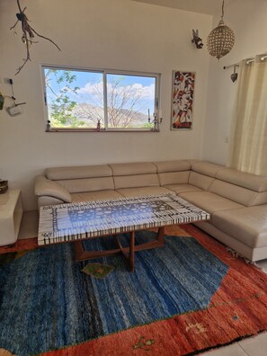 A living room with a beige sectional sofa, a patterned coffee table, a colorful rug, a large window with a view of trees and mountains, and a decorative hanging light fixture.