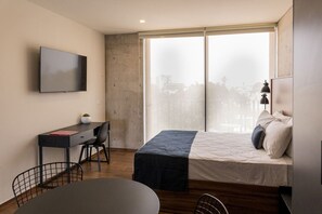 Ein modernes Schlafzimmer mit Bett, Schreibtisch, Stuhl, Fernseher und einem großen Fenster mit Blick auf die Stadt.