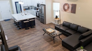 A modern living room with a black leather sofa, a coffee table, and a kitchen area with white cabinets and stainless steel appliances. - Steps to Beach Private Pool 2 Big Levels (South Padre Island)