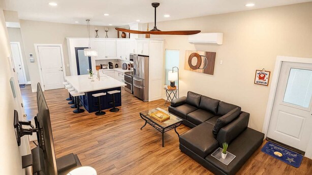 A modern living room with a sectional sofa, a coffee table, and a TV. The kitchen in the background has white cabinets and stainless steel appliances. - Steps to Beach Private Pool 2 Big Levels (South Padre Island)