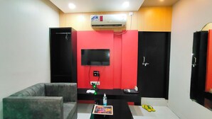 A hotel room with a grey sofa, a flat-screen TV mounted on a red wall, a small table with a notebook and a bottle, and a pair of yellow flip-flops on the floor. - Hotel Jay Deluxe (Sasaram)