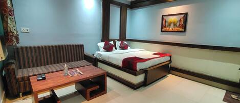 A compact hotel room with a sofa, a small table, a single bed, and a painting on the wall.