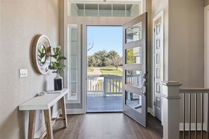 Un hall d’entrée moderne avec une porte vitrée donnant sur une terrasse, une console blanche ornée d’une plante en pot et un miroir fixé au mur.