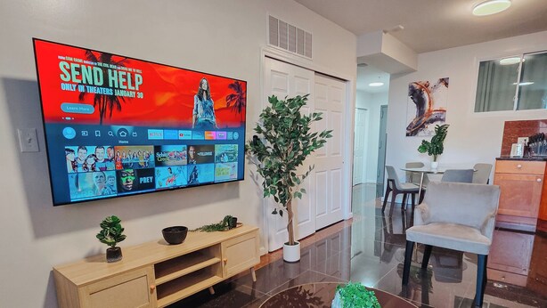 A modern living room with a flat-screen TV mounted on the wall, a wooden entertainment unit, a round coffee table, and a dining area with a chair and table. - Vale / King Bed / Elevator / Balcony / Ensuite (Philadelphia)
