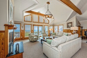 A spacious living room with a high ceiling, large windows, and a cozy fireplace. - Oceanfront, elevator, pool, hot tub, walk to shops. Friday check in. (Kitty Hawk)