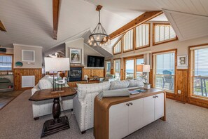 A spacious living area with a large sofa, a wooden coffee table, and a chandelier. There are multiple windows with blinds, a fireplace, and a television. - Oceanfront, elevator, pool, hot tub, walk to shops. Friday check in. (Kitty Hawk)