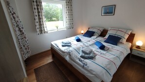 En snyggt bäddad säng med blått och vitt sänglinne, en sänglampa och ett fönster med gardiner.
