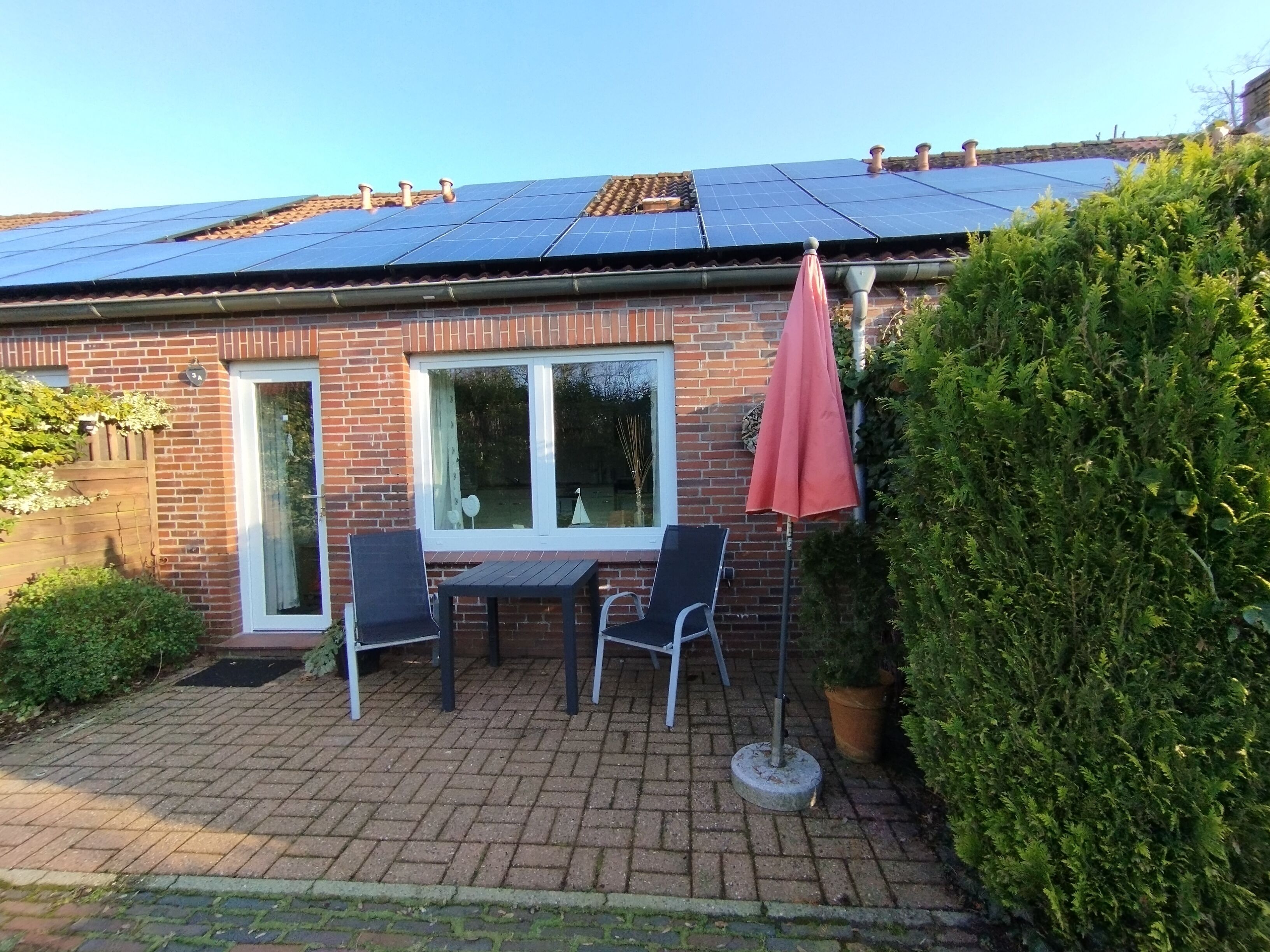 Een bakstenen huis met een dakbedekt met zonnepanelen, een terras met twee stoelen en een tafel, en een grote potplant.