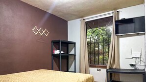 A bedroom with a bed, a small black shelf, a window with curtains, and a TV mounted on the wall. - Casa cómoda y tranquila a minutos de Xetulul (Guatemala)