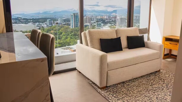 A modern living room with a sofa, a coffee table, and a view of a cityscape through large windows. - Cómodo apartamento a minutos de cayala (Guatemala City)