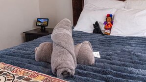 Ein Bett mit einer blauen Decke und einem gefalteten Handtuch. Es gibt einen kleinen Tisch mit einem Fernseher und einem dekorativen Kissen mit einer Spielzeugfigur.