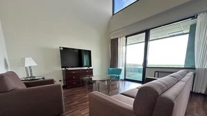Uma sala de estar moderna com um sofá, uma televisão, uma mesinha e vista para o mar através de grandes janelas.