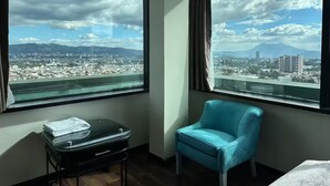 Quarto de hotel com uma grande janela que oferece vista para a cidade, uma poltrona azul e uma pequena mesa com uma toalha.