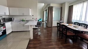 Cozinha moderna com armários brancos, micro-ondas e pia. Possui área de jantar com mesa e cadeiras, e vista para o exterior através de grandes janelas.