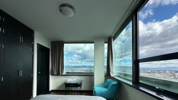 Quarto de hotel com uma cama grande, uma mesa pequena, uma cadeira azul e vista para o horizonte urbano através de grandes janelas.