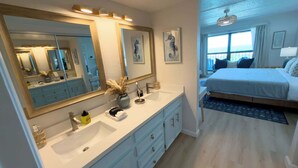 A modern bedroom with a large bed, a nightstand, and a bathroom with a double sink and a large mirror. - Saida 3603 - 6 Floor Gulf-front Remodel - Sleeps 8 (South Padre Island)