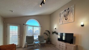 A living room with a sofa, a glass coffee table, a TV on a cabinet, a painting on the wall, and a window with blinds. - Calgary Home | 3BR Home | Sleeps 6 | Parking | 15min to Downtown (Calgary)