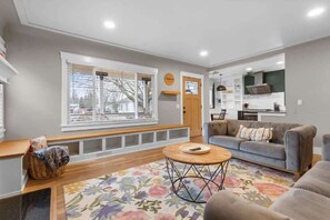 A modern living room with a grey sofa, a round coffee table, and a large window with blinds. - CDA Midtown Home, Near Lake & Downtown, Fire Pit (Coeur d'Alene)