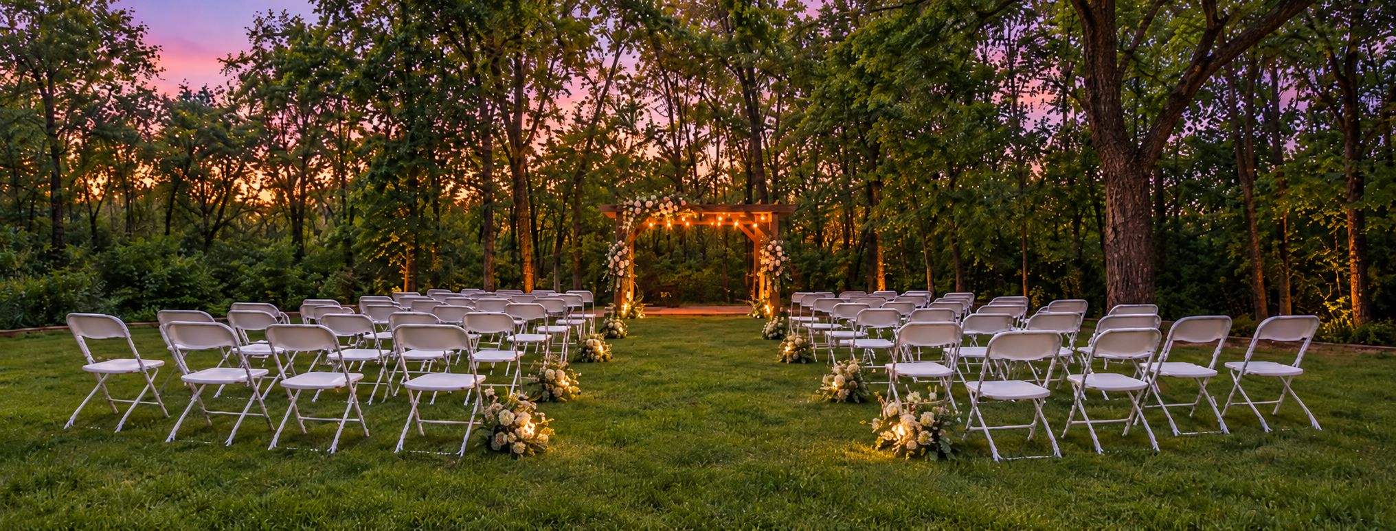 An outdoor wedding venue with rows of chairs arranged for guests, a floral archway, and a natural backdrop of trees.